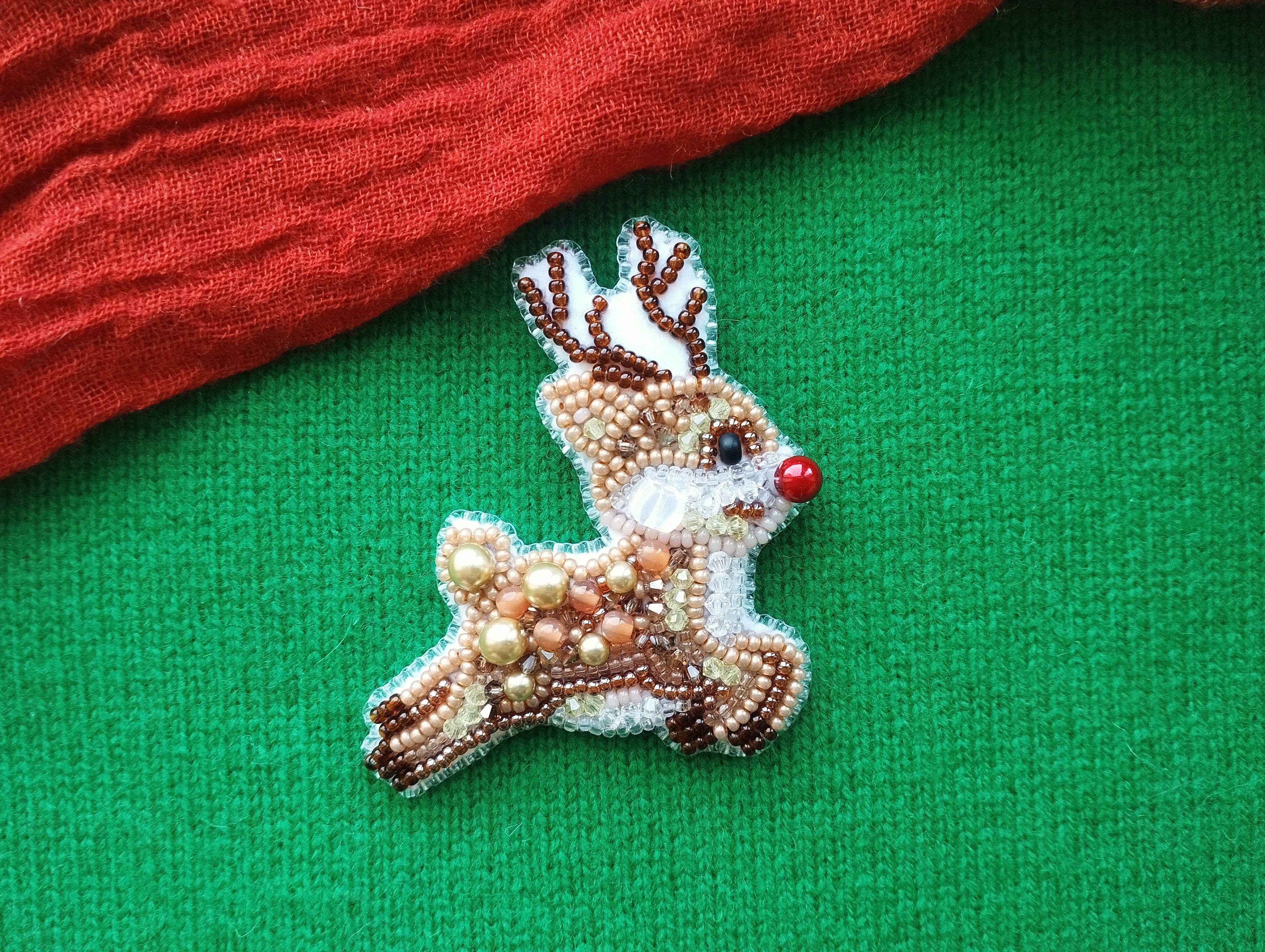 A beaded reindeer pin is displayed on a green fabric, with a red fabric piece partially visible to the left.